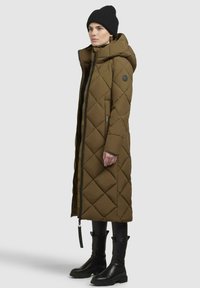 Olive green, long puffer coat with diamond quilting, high collar, and a zippered front. Worn with black pants and ankle boots, accessorized with a black beanie.