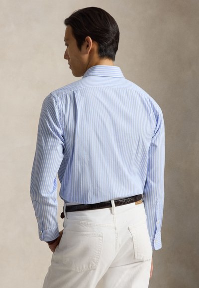 Mann trägt ein hellblau gestreiftes Langarmhemd, in weiße Hose mit dunklem Gürtel gesteckt, steht mit dem Rücken zu einem einfarbigen Hintergrund.