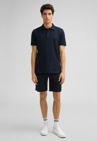 Marineblaues Poloshirt mit kurzen Ärmeln und dreiknöpfigem Kragen, kombiniert mit schwarzen Shorts. Weiße Slipper und kontrastierende Crew-Socken.