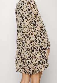 Robe en imprimé léopard en tissu léger et fluide. Dotée de manches longues, d'une jupe à volants et d'un col haut. Les couleurs incluent beige, marron et noir.