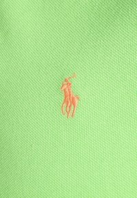 Grön pikétröja i tyg med texturerad stickning. Har en orange broderad logotyp av en häst och ryttare. Enkel kragsdesign.