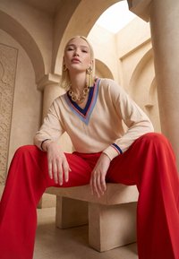 Femme blonde portant un pull crème avec des bordures bleues et rouges, un pantalon large rouge, des boucles d'oreilles dorées en forme de créoles et un collier doré épais, assise dans un intérieur voûté.