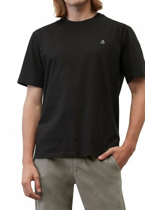 Junge Person trägt ein schlichtes schwarzes T-Shirt mit kleinem weißem "M O P" Logo auf der Brust und hellgraue Hose, Hand in der Tasche.