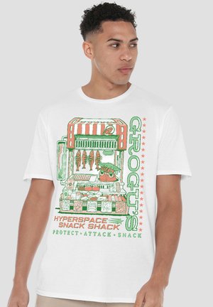 Junger Mann in weißem T-Shirt mit farbenfroher Grafik eines Imbissstandes, die Fisch, Gemüse und den Text „Grog's Hyperspace Snack Shack“ zeigt.