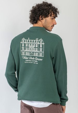 Uomo con capelli ricci che indossa una camicia verde a maniche lunghe con un’illustrazione bianca di un edificio e testo sul retro, di profilo su uno sfondo semplice.