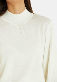 Witte geribbelede gebreide trui met een hoge hals, voorzien van een geplooide detail en kleine decoratieve parels langs de kraag.