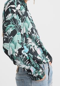 Donna che indossa una blusa nera a maniche lunghe con stampa botanica verde e bianca, infilata in jeans blu, con le mani nelle tasche.