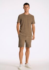 T-shirt in maglia marrone e pantaloni corti abbinati, maniche corte, colletto girocollo, texture morbida, modello in piedi con una mano nella tasca dei pantaloni corti, sneakers bianche.