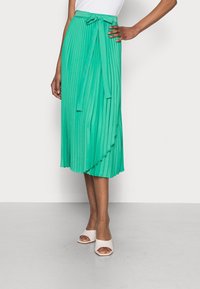 Gonna midi plissettata di un vivace verde con un dettaglio in vita legato. Tessuto morbido e design fluido, completato da morbide scarpe bianche con punta aperta.