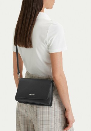 Kobieta o prostych brązowych włosach nosi białą koszulę z krótkim rękawem i spodnie w kratę, nosząc czarną torebkę na ramię marki Lasocki z klapką.