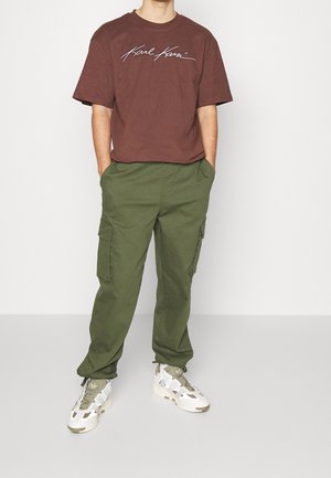 Camiseta de algodón marrón con logo en escritura blanca, combinada con pantalones cargo verde oliva con bolsillos laterales y un corte relajado. Zapatillas blancas.