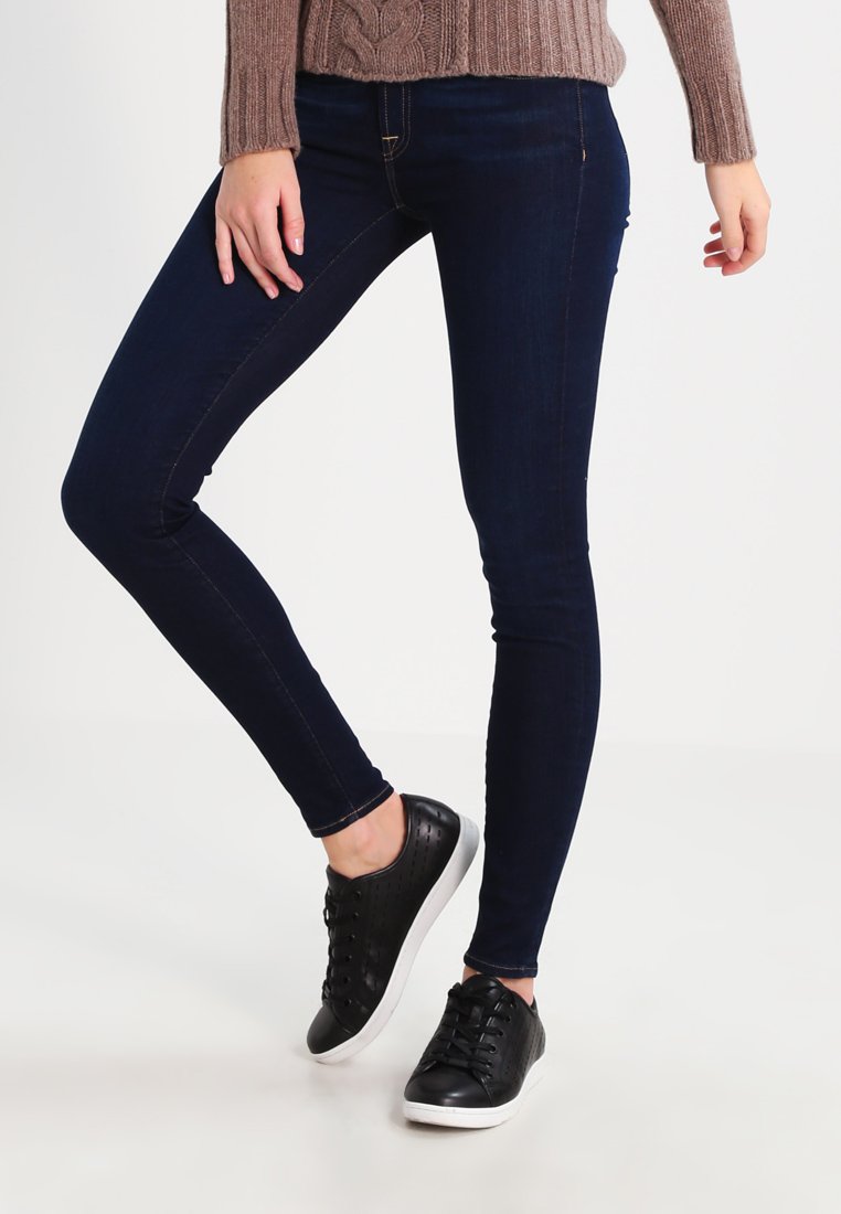 Fatos de ganga skinny azul escuro com um tecido de ganga elástico, apresentando uma cintura alta e detalhes de costura discretos, combinados com ténis pretos.