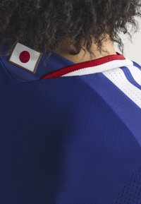 Blå strukturerad sporttröja med röda och vita accenter. Har en liten japansk flagga broderad på axeln. Närbild.