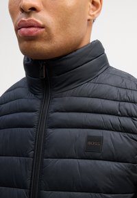 Μαύρο padded μπουφάν με ψηλό γιακά, διαθέτει ορατό φερμουάρ και τετράγωνη μαύρη ετικέτα λογότυπου που γράφει "BOSS" στο στήθος.