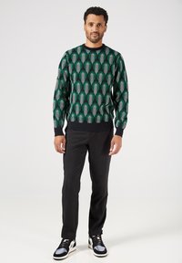 Maglione a maglia verde con motivo a archi bianchi, abbinato a pantaloni neri e sneakers multicolori.