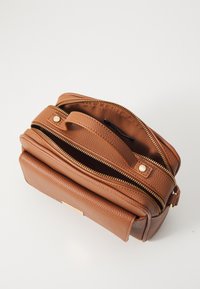 Sac à main en cuir marron avec fermeture éclair, finition texturée, compartiments doubles et une poignée supérieure. Détails de broderie et accents dorés visibles.