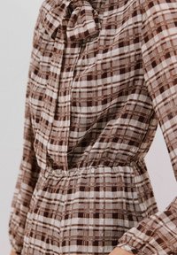 Braun- und cremefarbener Karo-Bluse mit einer Schleife am Halsauschnitt, langen Ärmeln, gerafftem Bund und einem leichten, strukturierten Stoff.