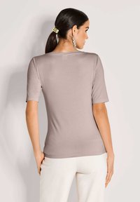 Vrouw met donker haar in een paardenstaart draagt een taupe top met korte mouwen en witte broek, met haar rug naar de camera, tegen een effen lichte achtergrond.