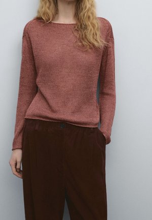 Femme portant un pull tricoté rose pâle à manches longues et un pantalon taille haute marron foncé, debout devant un fond gris clair uni.