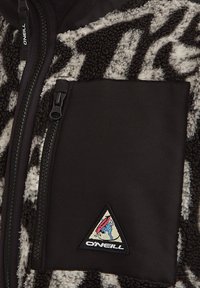 Pull en polaire avec un motif noir et blanc, fermeture éclair à l'avant, grande poche poitrine et un patch de logo triangulaire dans le coin.