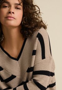 Beige V-Ausschnitt-Pullover mit dunkelblauen horizontalen Streifen. Gerippte Struktur, weite Ärmel und lockere Passform. Fokus auf Oberkörper und Ausschnitt.