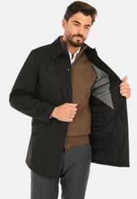 Cappotto esterno nero con colletto, chiusura con bottoni e due tasche. Maglione marrone a strati sotto. Interno foderato mostra dettagli in tessuto grigio.