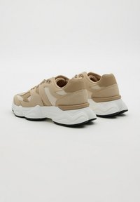 Zapatillas deportivas gruesas en beige y blanco con paneles de ante y malla, suelas gruesas blancas y taco negro, vistas desde la parte trasera sobre un fondo blanco.