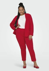 Sacou roșu și pantaloni tapered asortați cu o cămașă albă. Textură asemănătoare satinului cu un finisaj neted. Pantofi cu vârf închis roșii completează ținuta.