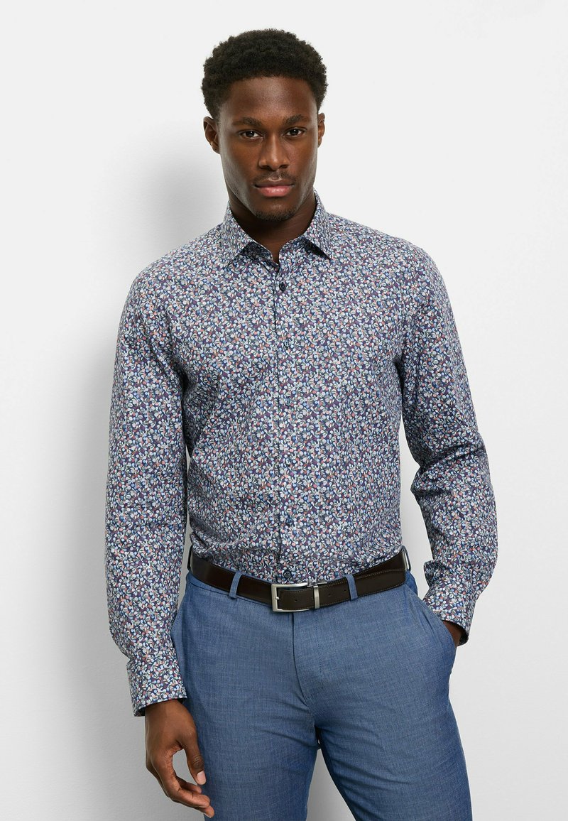 Langärmliges Hemd mit floralem Muster auf blauem Hintergrund, mit mehrfarbigen Blattmustern, einem normalen Kragen und einer geknöpften Vorderseite. Kombiniert mit hellblauen Hosen.