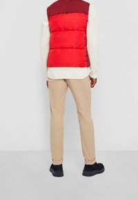 Hombre con chaleco acolchado rojo y burdeos, suéter de punto blanco, pantalones beige y zapatos negros de pie, con la espalda hacia la cámara.