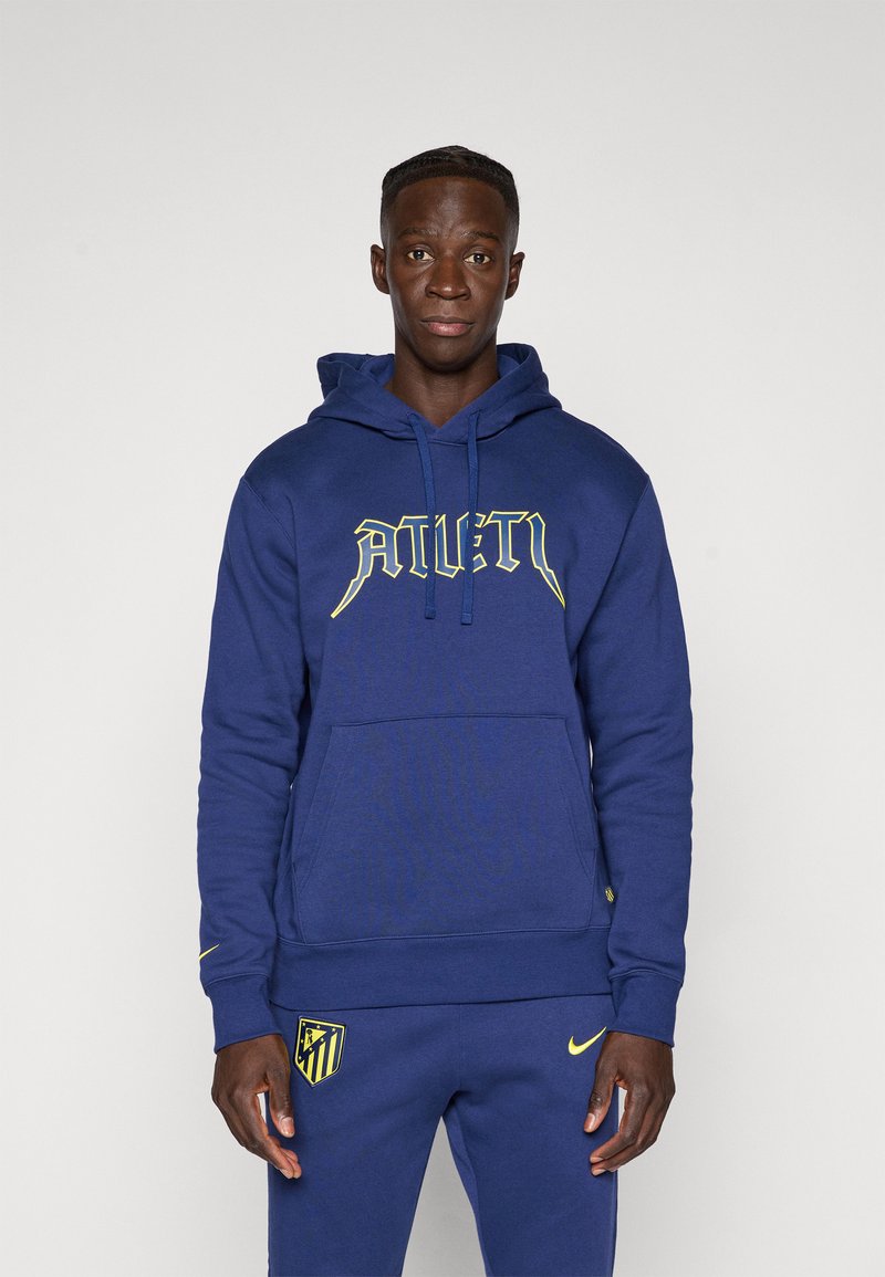 Mand iført en blå Atletico Madrid hoodie og sweatpants med gule logoer, stående foran en ensfarvet lys baggrund.