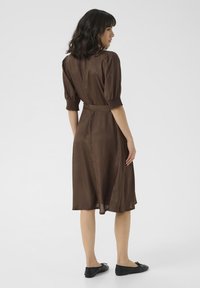 Robe brune midi avec des manches smockées et une taille cintrée. Tissu lisse et design fluide. Des chaussures plates noires complètent la tenue.