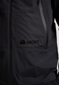 Svart jacka med texturerat, lättviktigt tyg. Har en ficka med dragkedja och präglad "JACK1T"-logotyp på vänster sida.