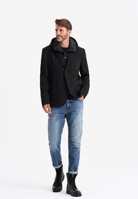 Svart skräddarsydd jacka med huva, i kombination med en svart skjorta, ljusblå slitna jeans och svarta snörboots.