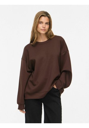 Ung kvinde iført en løs brun sweatshirt og sorte bukser, stående med den ene hånd i lommen mod en ensfarvet hvid baggrund.