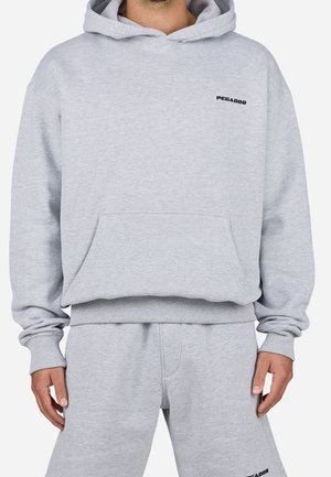 Homme portant un sweat à capuche gris clair et un pantalon de survêtement assorti avec de petits logos noirs "PEGADOR" sur la poitrine et la cuisse.