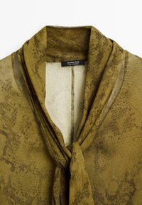 Blouse en tissu imprimé serpent vert olive avec un nœud noué au col et une étiquette de marque Massimo Dutti visible à l'intérieur du col.