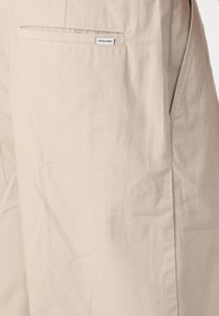 Shorts bej dintr-un material ușor, cu un buzunar la spate care prezintă o etichetă și o textură netedă cu cute subtile.