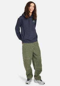 Marineblaues Kapuzensweatshirt mit Fronttasche, kombiniert mit übergroßen olivgrünen Hosen mit genähten Paneelen. Sportliche Freizeitschuhe runden den Look ab.