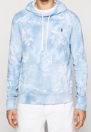 Jasnoniebieska bluza z kapturem w stylu tie-dye z białymi hawajskimi motywami kwiatowymi i krajobrazowymi, białymi sznurkami oraz kieszenią typu kangur, noszona z białymi spodniami.