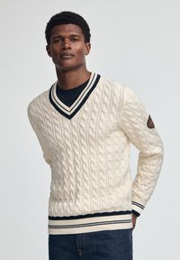 Kremowy sweter w splot warkoczowy z dekoltem w kształcie litery V, czarno-białym kołnierzykiem w paski oraz ściągaczem w pasie. Posiada naszywkę z logo na lewym rękawie.