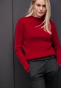 Rode geribde trui met een hoge hals, lange mouwen en witte zijnaden, gedragen met grijze tailor-made broeken met voorzakken.
