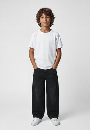 Niño con cabello rizado está de pie con las manos en los bolsillos, lleva una camiseta blanca, vaqueros negros anchos y zapatillas blancas sobre un fondo liso.