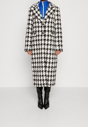 Person iført en sort og hvid hundetand-lang frakke, blå lynlås-trøje og sorte højhælede støvler, stående foran en ensfarvet baggrund.