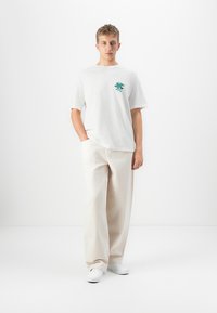 Ung mand står med hænderne i lommerne, iført hvid t-shirt med grafisk tryk, beige vide bukser og hvide sneakers foran en ensfarvet baggrund.