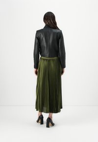 Veste en cuir noir courte associée à une jupe plissée olive jusqu'à la cheville, présentant une texture lisse et des chaussures à talons hauts noires.
