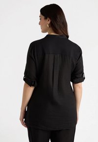 Camisa negra de tejido ligero con mangas enrolladas y un panel trasero sólido en contraste. Presenta un cuello y un diseño de ajuste relajado.