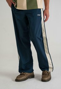 Pantaloni largi bleumarin cu dungi crem pe laterale, confecționați dintr-un material ușor și având un mic logo pe coapsa stângă.