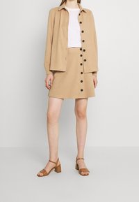 Giacca beige con un design strutturato, abbinata a una gonna beige coordinata con bottoni neri. Sandali con tacco a blocco marroni completano il look.