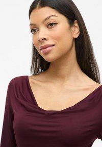 Vrouw met lang, donker haar die een diep paarse bootcut-top draagt, lichtjes opzij kijkend met een neutrale uitdrukking tegen een witte achtergrond.